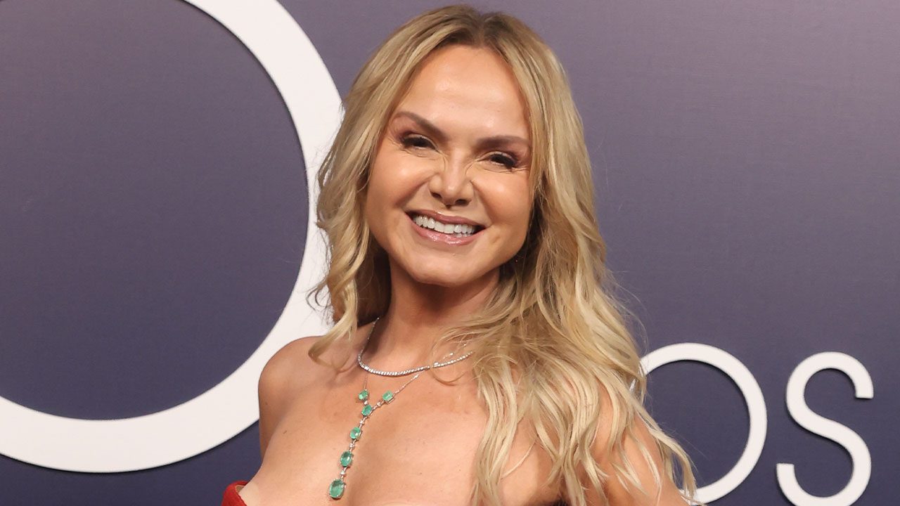 Eliana sorrindo em evento com vestido vermelho e colar de esmeraldas diante de painel com logotipo circular