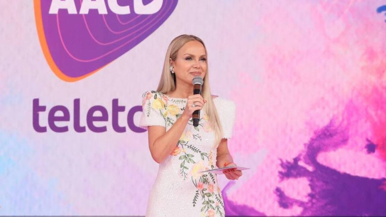 Eliana de vestido branco com bordados florais segura microfone no palco do Teleton diante de painel com logotipo da AACD