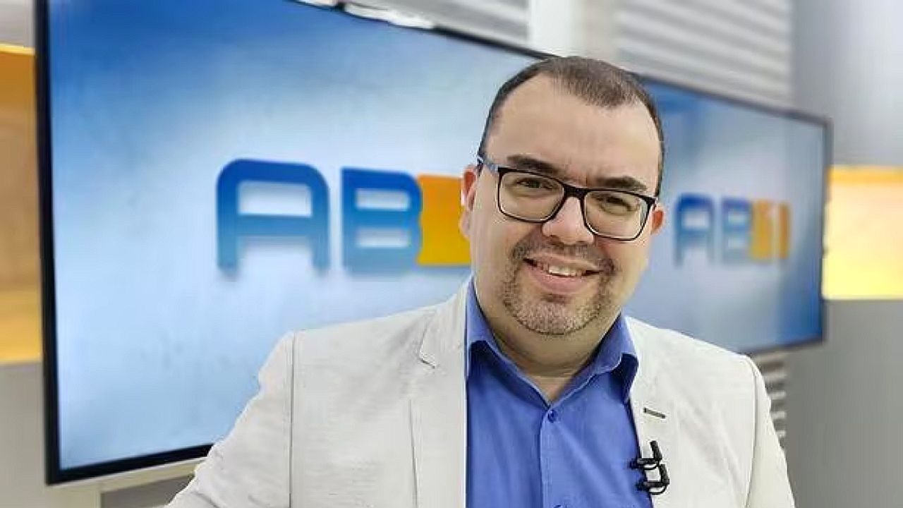 Apresentador sorri em estúdio do AB1 diante de telão com logotipo do telejornal da TV Asa Branca
