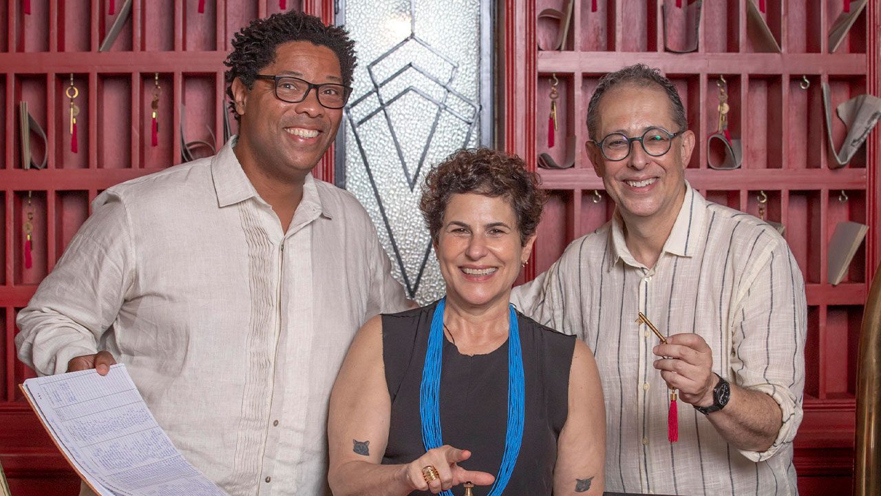 Elisio Lopes Jr., Duca Rachid e Júlio Fisher posam sorrindo com chave de hotel em frente a painel vermelho decorado