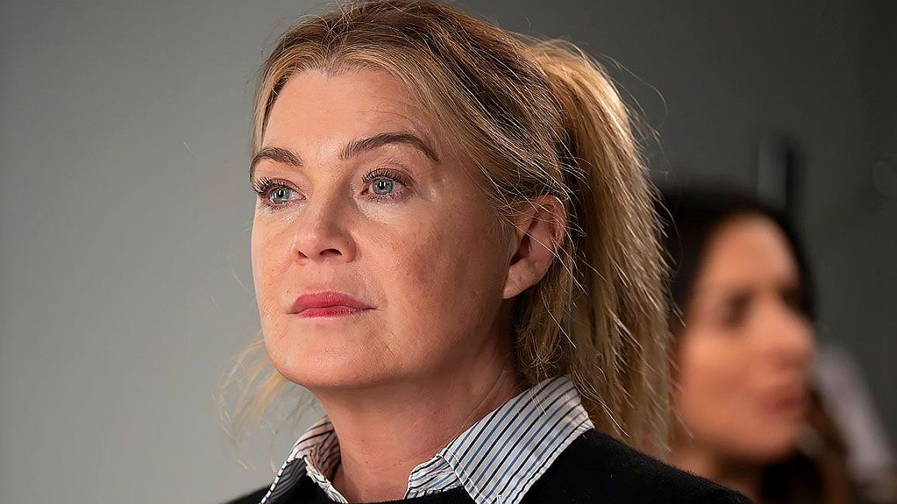 Mulher loira de cabelo preso observa algo à frente com expressão séria e reflexiva em ambiente interno com pessoa desfocada ao fundo