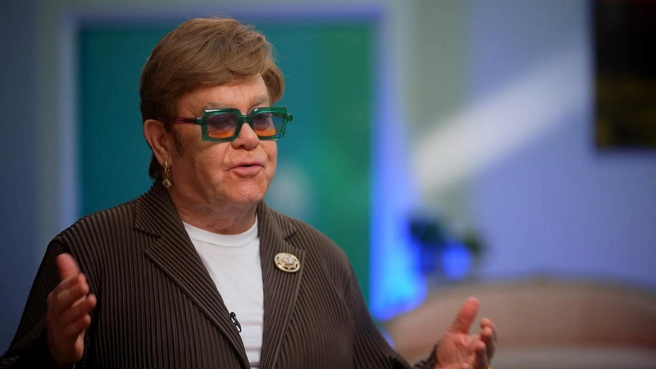 Pessoa idosa de pele clara aparece em plano médio em estúdio de televisão, com cabelo curto castanho grisalho bem penteado, usando óculos de armação grossa em tom verde translúcido com lentes levemente coloridas, expressão séria e concentrada com boca entreaberta como se estivesse falando, vestindo blazer marrom de risca fina com broche dourado na lapela esquerda e camiseta branca por baixo