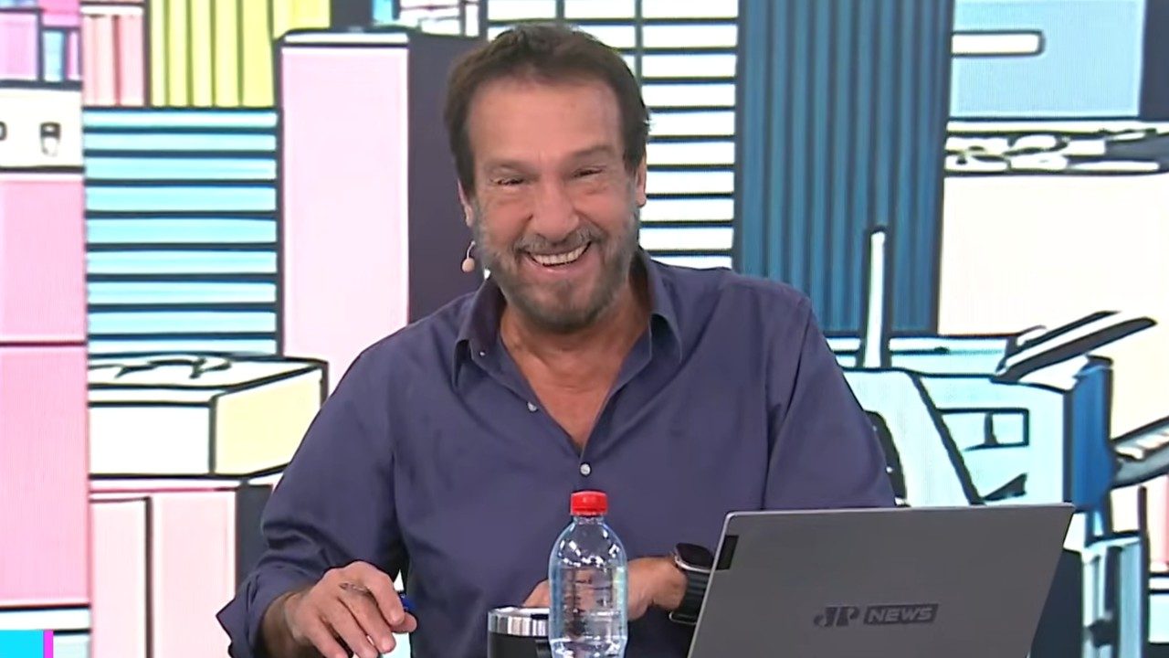 Emílio Surita sorri à bancada com camisa azul diante de notebook e garrafa de água em estúdio da Jovem Pan News