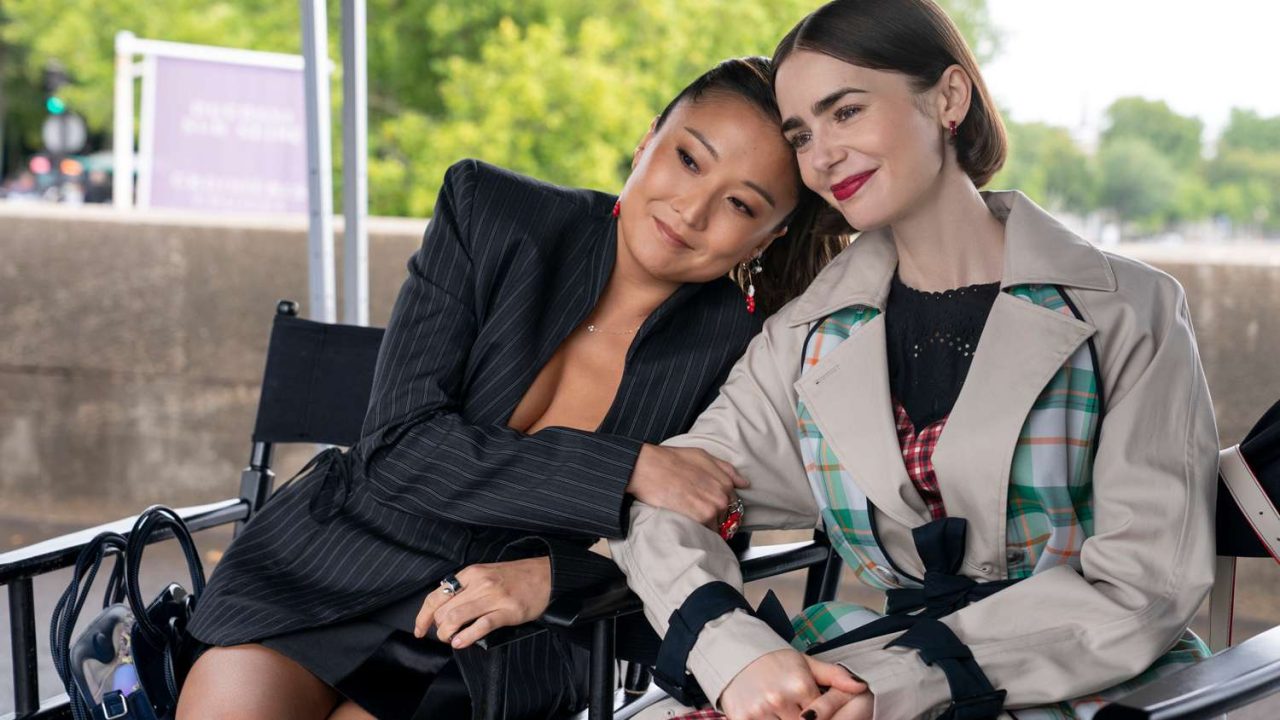 Emily Cooper e Mindy Chen sentadas em cadeiras de diretor apoiam a cabeça uma na outra em bastidores ao ar livre da série Emily in Paris