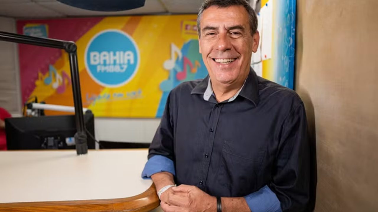Emmerson José no estúdio da Rádio Bahia FM, sorrindo próximo ao microfone e banner da emissora