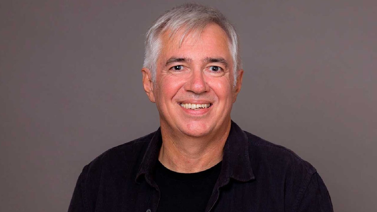 A imagem mostra um homem sorridente, de cabelos grisalhos curtos, vestindo uma camisa escura sobre uma camiseta preta, posando contra um fundo neutro. A expressão é simpática e transmite naturalidade