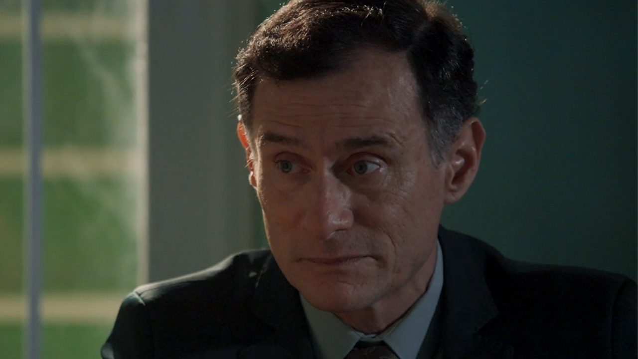 Homem de terno escuro olha para o lado com expressão séria em ambiente interno iluminado por luz natural