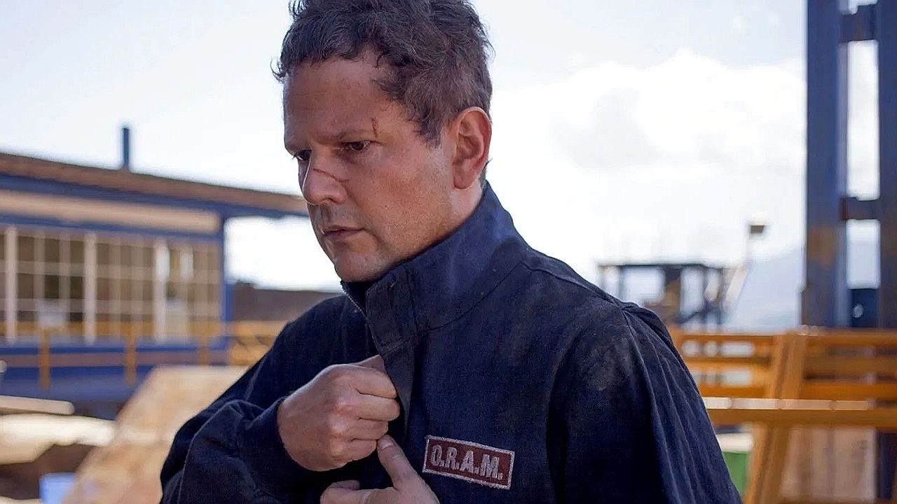 Homem com uniforme escrito Q R A M ajusta gola do casaco em área industrial ao ar livre