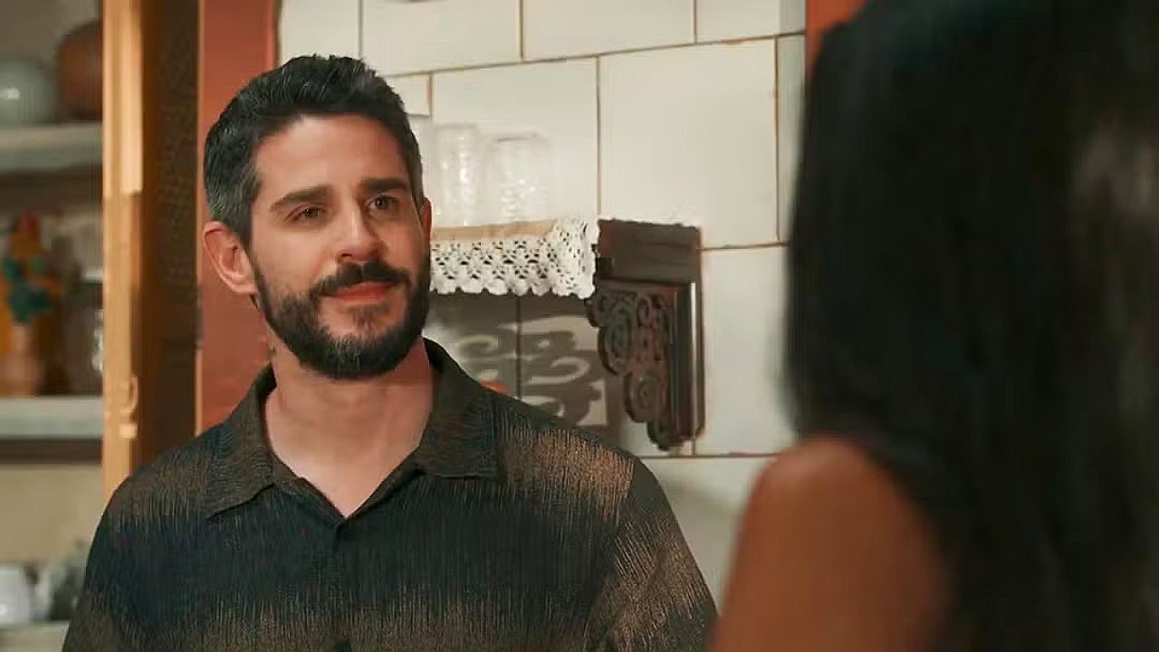 Homem com barba e cabelo curto escuro conversa com alguém em uma cozinha, usando camisa escura, enquanto olha com expressão calma para uma mulher que aparece de costas no primeiro plano