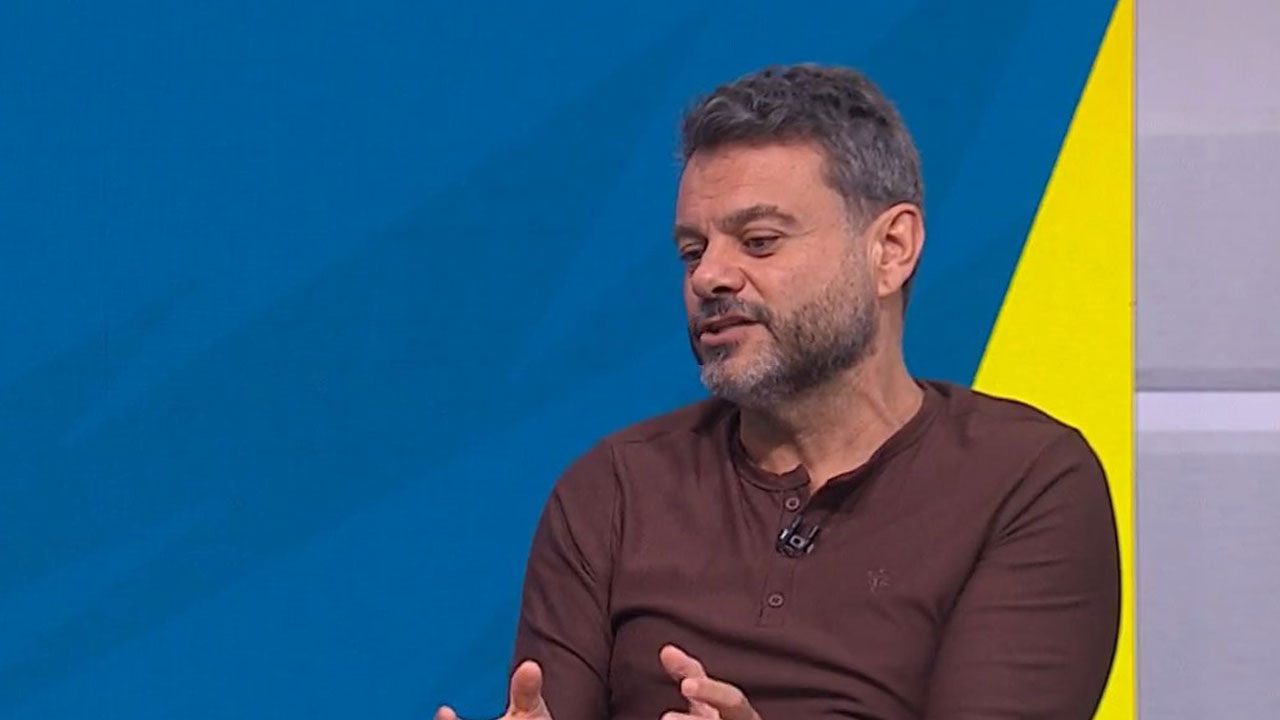 Eric Faria sentado e gesticulando durante participação em programa esportivo com fundo azul e amarelo