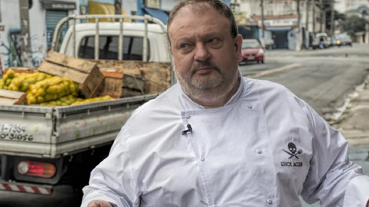 Homem com jaleco de chef branco e logotipo bordado caminha em rua com caminhão de frutas ao fundo