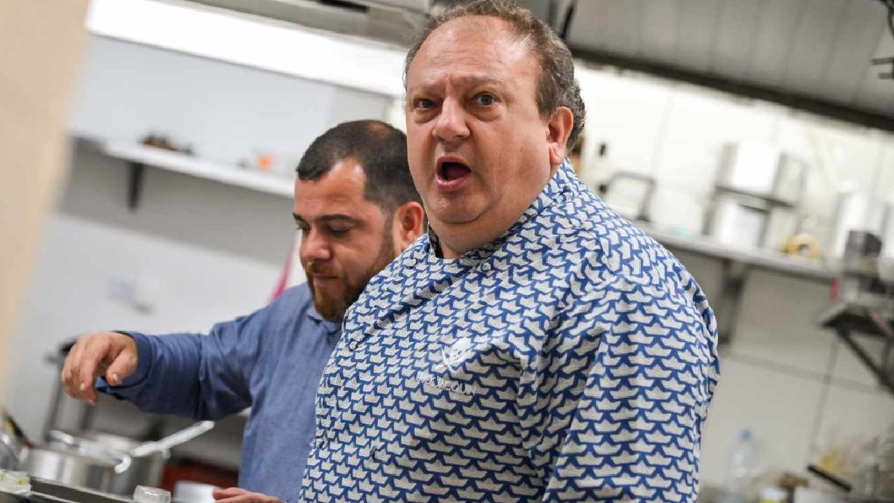 Homem com expressão surpresa em cozinha profissional enquanto outro cozinheiro trabalha ao fundo durante cena de programa ou filme gastronômico