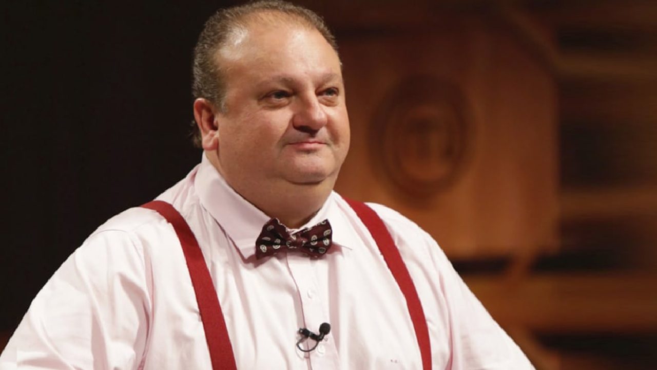 Erick Jacquin em um estúdio de programa culinário, usando camisa social rosa, gravata borboleta estampada e suspensórios vermelhos, com expressão séria e o logotipo do MasterChef desfocado ao fundo