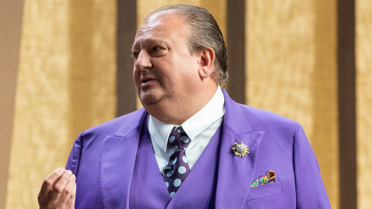 A imagem mostra Erick Jacquin de perfil em um estúdio, usando um terno roxo chamativo com gravata de bolinhas e lenço colorido no bolso. Ele faz um gesto com a mão enquanto fala, enquanto apresenta o MasterChef Celebridades. O fundo é composto por tons quentes e padrões verticais.