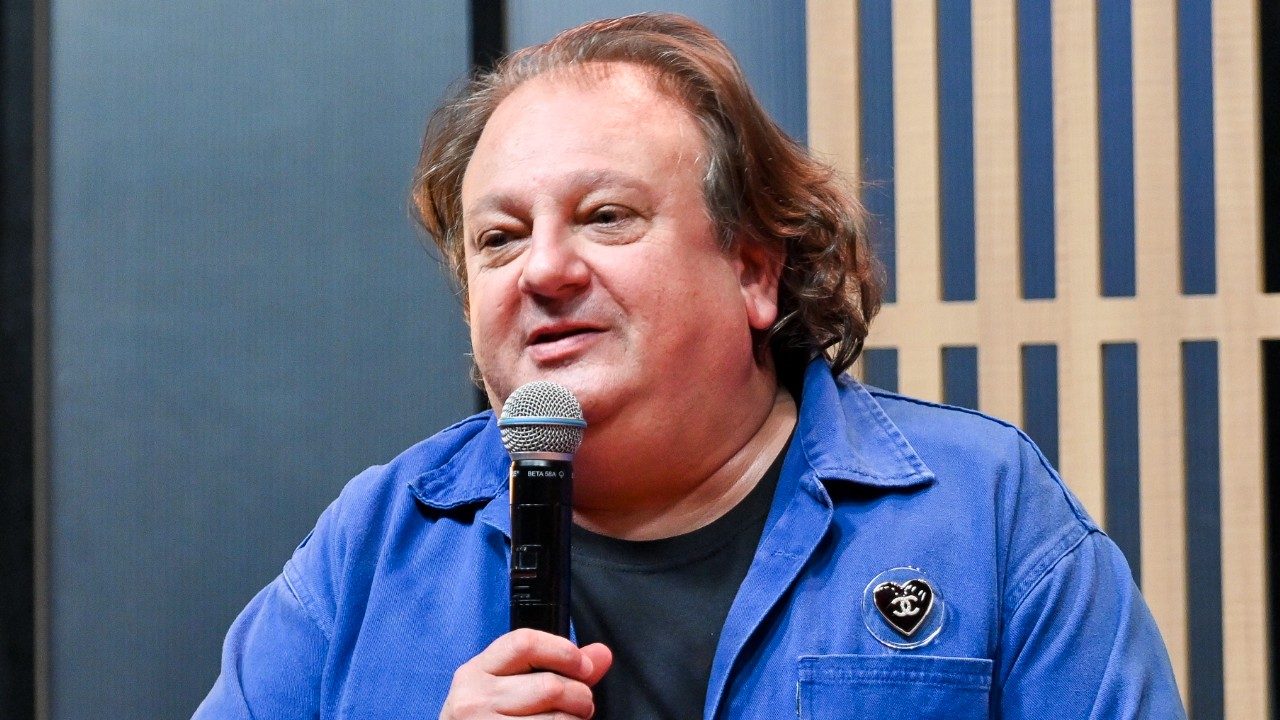 Erick Jacquin de jaqueta azul segura microfone e fala com leve sorriso diante de painel com ripas verticais de madeira