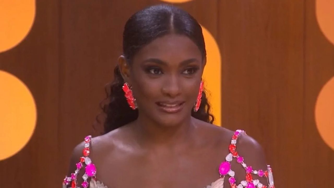 Mulher com cabelo preso para trás usa vestido claro adornado com pedrarias rosas e vermelhas. Ela também usa brincos grandes combinando com o look. A cena ocorre em um estúdio iluminado com luz quente e círculos amarelos decorativos ao fundo.