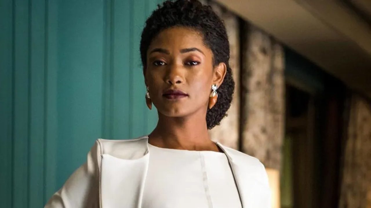 Mulher negra com expressão séria, usando vestido e blazer brancos e brincos elegantes, posa em ambiente interno com parede verde e detalhes em madeira ao fundo.