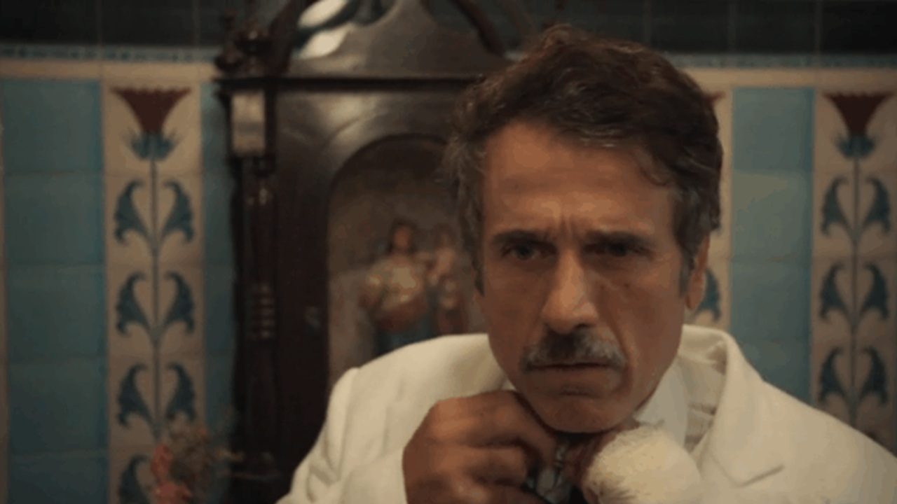 Ernesto ajusta a gravata com expressão séria em cena da novela Êta Mundo Melhor! da TV Globo
