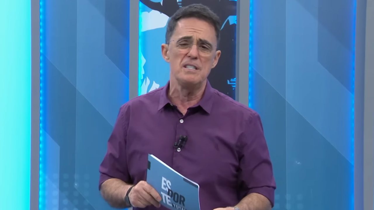 Fernando Fernandes, apresentador com camisa roxa, segura cartão do Esporte Total no estúdio da Band, com telão azul ao fundo.