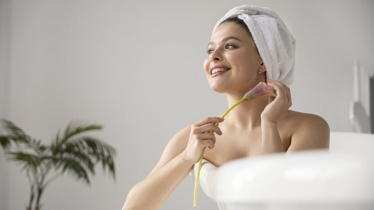 Mulher com toalha na cabeça segura flor e sorri em ambiente claro sugerindo rotina de cuidados com a pele em casa