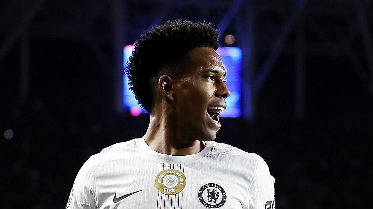 Jogador Estêvão com expressão de alegria e boca aberta veste camisa branca do Chelsea com escudos em destaque, posicionado em campo de futebol à noite