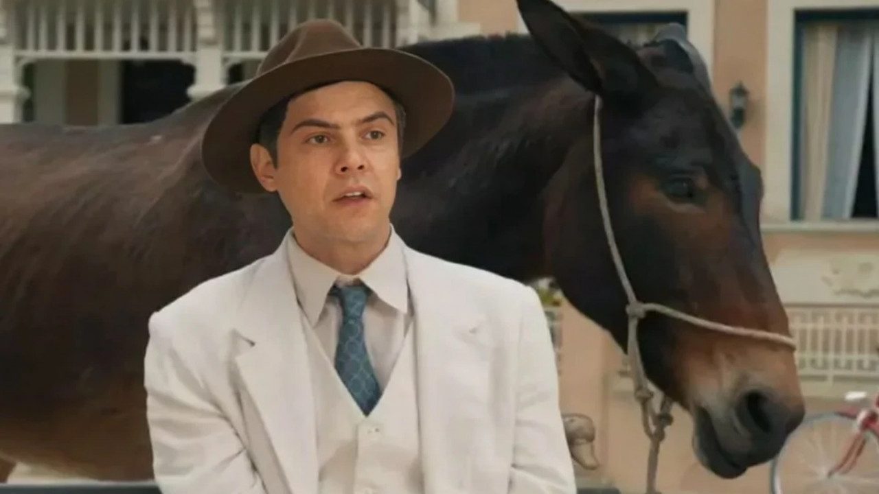 A imagem mostra um homem vestindo terno branco, gravata azul e chapéu marrom, em frente a um prédio de fachada clara com janelas abertas. Atrás dele, há uma mula marrom presa por uma corda. A cena parece ambientada em uma cidade pequena, evocando um clima de época