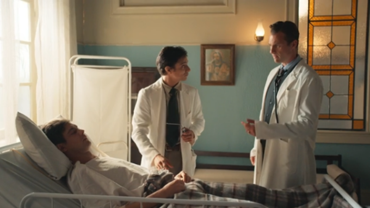 Cena em um quarto de hospital antigo, iluminado pela luz natural da janela. Um paciente jovem repousa em uma cama inclinada, coberto por um cobertor xadrez. Ao lado dele, dois médicos de jaleco branco conversam enquanto avaliam seu estado; um deles segura um instrumento médico. O ambiente tem paredes em tom pastel, vitrais decorativos e poucos móveis, reforçando a ambientação de época