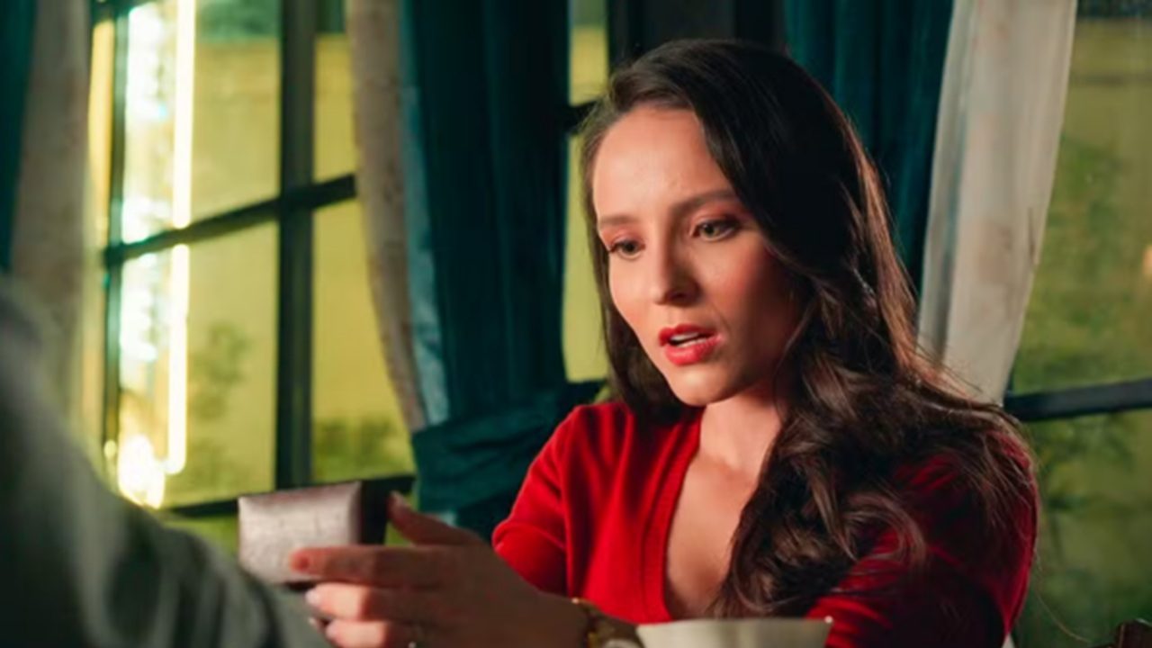 Mulher de cabelos castanhos longos e blusa vermelha observa surpresa uma pequena caixa que segura nas mãos, em ambiente interno com janelas grandes e cortinas azul-escuras ao fundo.