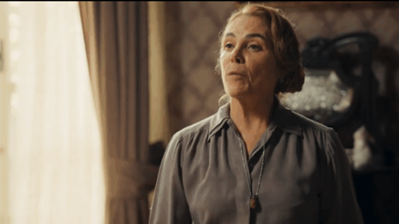 Mulher de expressão séria está em um ambiente interno com cortinas claras e papel de parede antigo, vestindo blusa cinza e colar simples, transmitindo tom de tensão ou reflexão