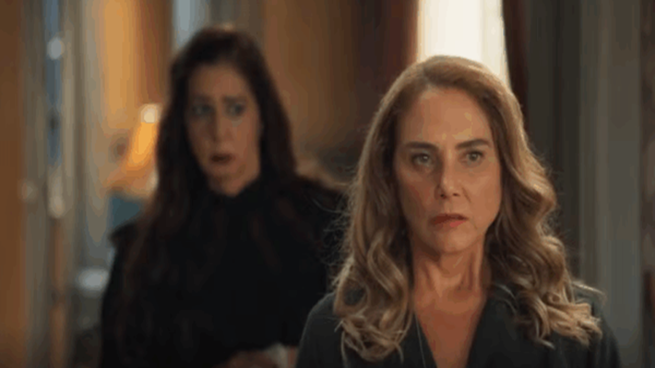 Cena mostra duas mulheres em um ambiente interno bem iluminado. A mulher em primeiro plano, de cabelos loiros ondulados, tem expressão séria e olhar fixo à frente, enquanto a mulher ao fundo, de cabelos castanhos escuros, parece surpresa ou preocupada