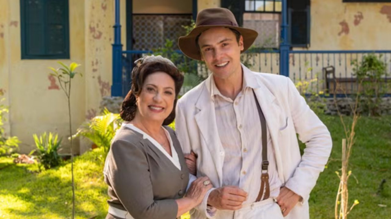 Uma mulher e um homem posam sorrindo em um jardim, em frente a uma casa antiga. A mulher veste um traje tradicional em tons de cinza e branco, enquanto o homem usa roupas claras, incluindo um chapéu marrom e suspensórios. Eles aparecem de braços dados, transmitindo proximidade e simpatia.