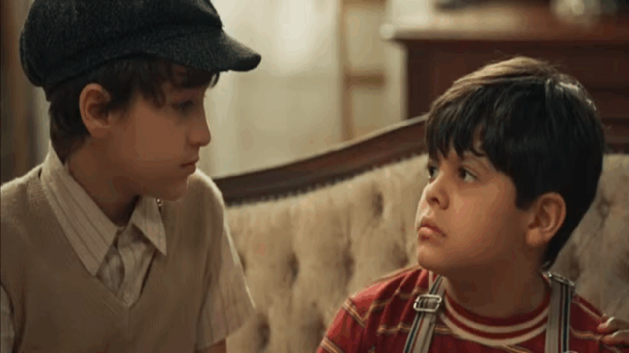 Dois meninos sentados em sofá antigo trocam olhares sérios em cena de época da novela Êta Mundo Melhor! da Globo