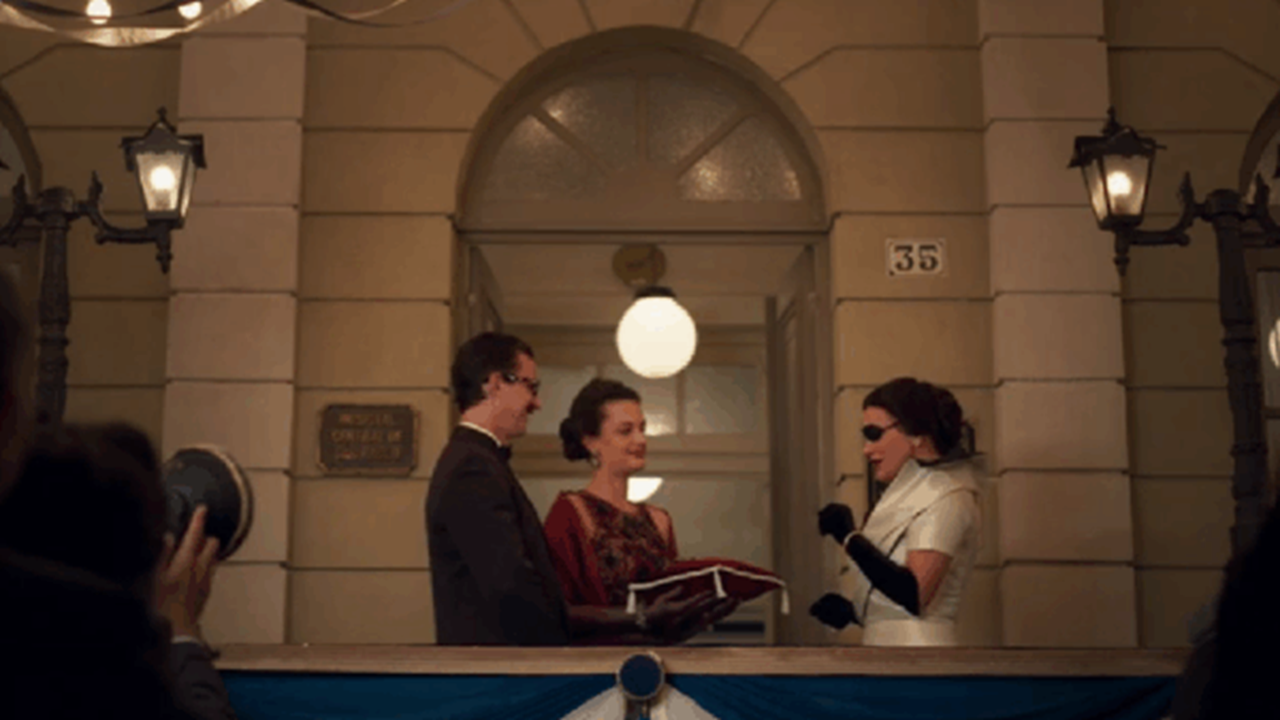 Cena de época mostra três pessoas em uma varanda elegante, diante de um prédio com fachada clássica e iluminação suave. Uma mulher com vestido branco, luvas pretas e óculos escuros assina um livro segurado por outra mulher, enquanto um homem de terno observa. Fotógrafos registram o momento, sugerindo um evento oficial ou cerimônia pública.