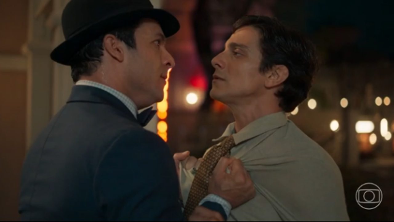 Dois homens adultos se encaram de perto em uma rua iluminada à noite. Um deles, usando terno escuro e chapéu, segura o outro pelo colarinho e pela gravata. O segundo homem veste camisa clara e expressão tensa. Ao fundo, luzes desfocadas da rua reforçam o clima de confronto e tensão da cena.