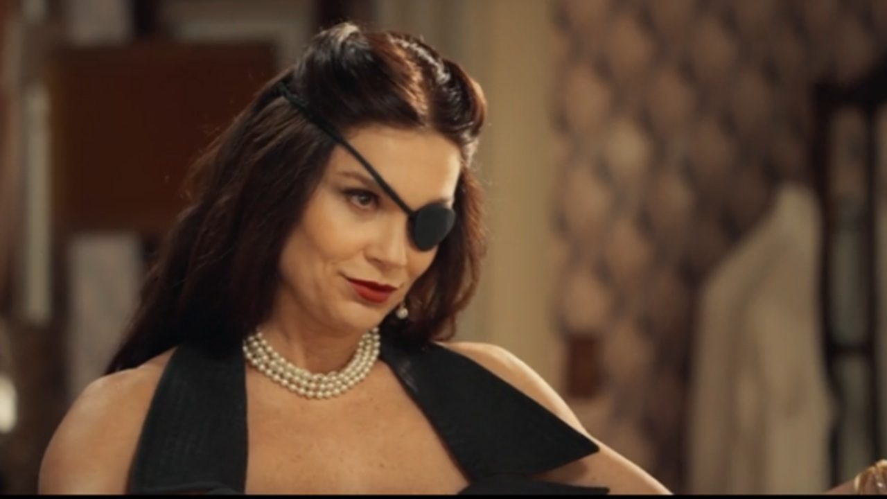 Mulher com tapa-olho preto no olho direito aparece em ambiente interno, usando vestido preto sem mangas e colar de pérolas. Ela tem cabelos castanhos presos parcialmente para trás, maquiagem marcada e expressão confiante, olhando levemente para baixo. Ao fundo, há móveis e papel de parede decorado, sugerindo um cenário elegante ou de época.