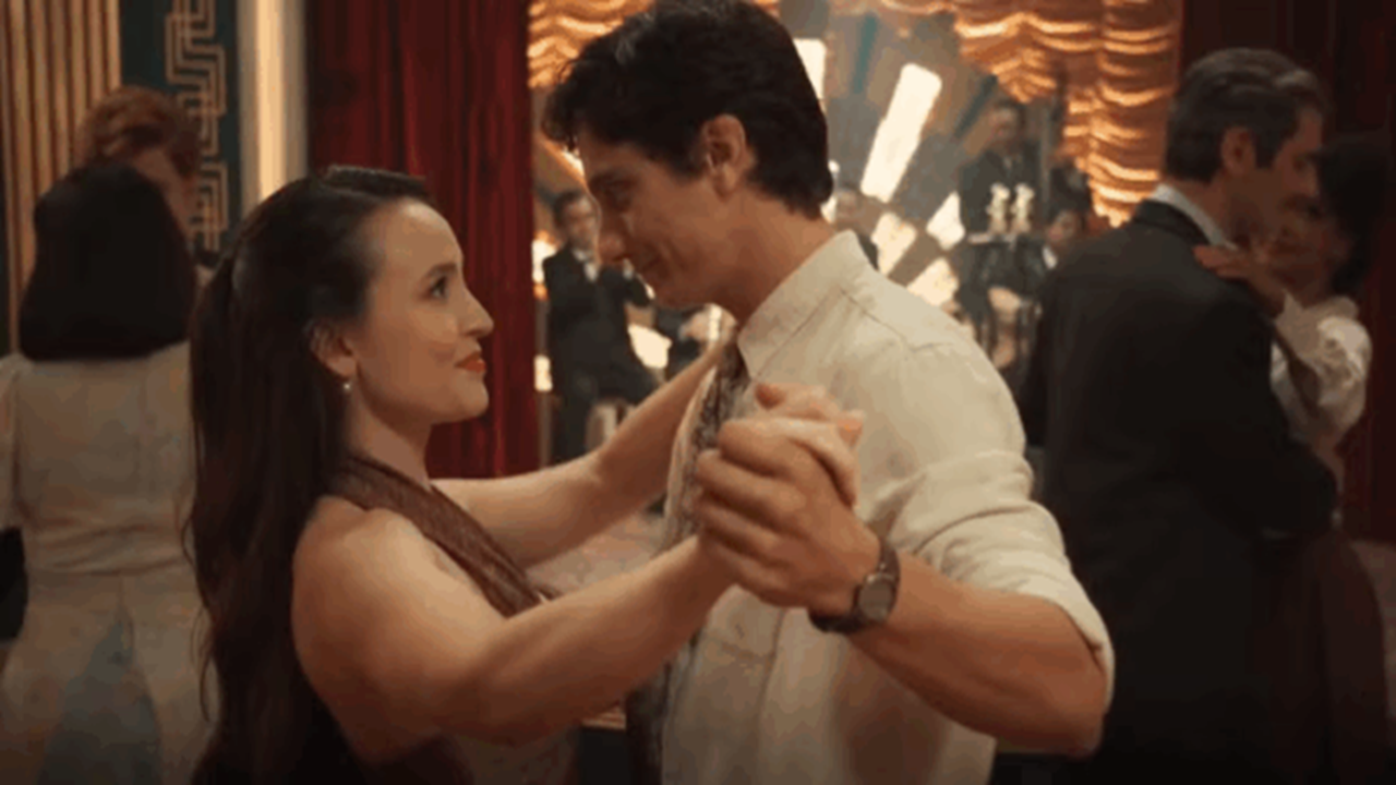 A imagem mostra um casal dançando em um salão elegante, com cortinas vermelhas e luzes douradas ao fundo. Ambos se olham com ternura enquanto seguram as mãos. Outras pessoas também dançam ao redor, criando um clima de romance e nostalgia