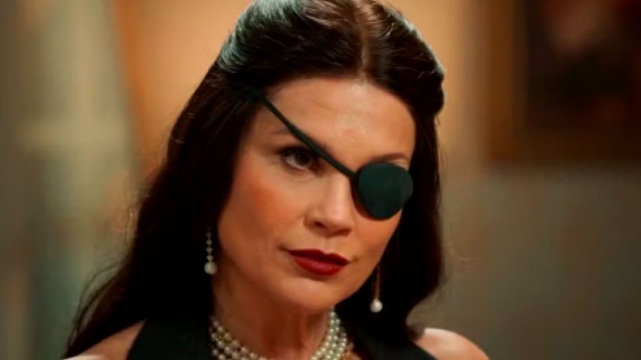 Mulher de cabelos escuros usa um tapa-olho e brincos de pérola, com expressão firme e olhar intenso, em ambiente interno com iluminação quente e fundo desfocado.