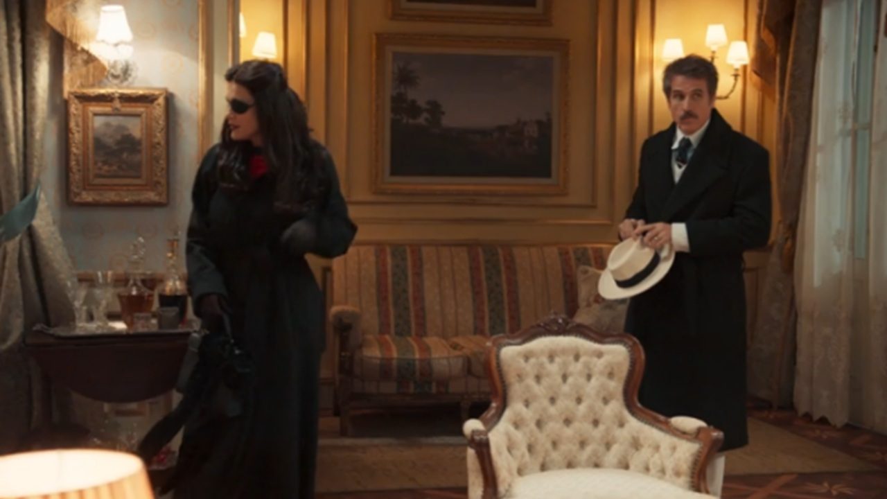 Homem de bigode usando sobretudo escuro e segurando chapéu branco observa mulher de cabelos escuros vestida com casaco preto e óculos escuros em sala elegante decorada com cortinas pesadas, poltronas estofadas, quadros na parede e iluminação amarelada de abajures e arandelas