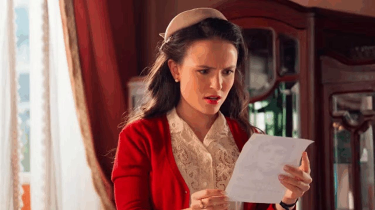 Cena da novela Êta Mundo Melhor!, mostrando uma mulher com expressão surpresa ou preocupada enquanto lê uma carta ou documento, em um ambiente de época com cortinas e móveis antigos ao fundo