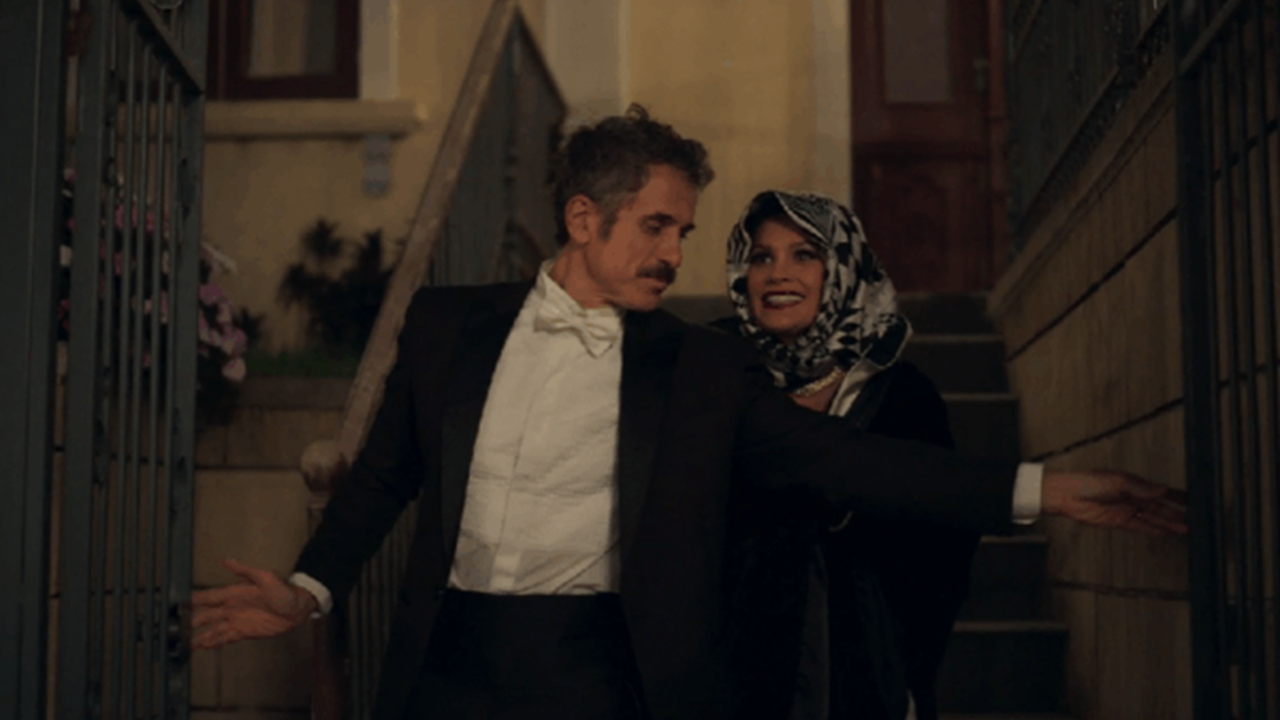 A imagem mostra um homem e uma mulher descendo uma escadaria externa à noite. O homem, de terno preto e camisa branca com gravata borboleta, faz um gesto expansivo com os braços enquanto sorri. A mulher, que usa um lenço estampado cobrindo a cabeça e um casaco escuro, o acompanha com um olhar alegre