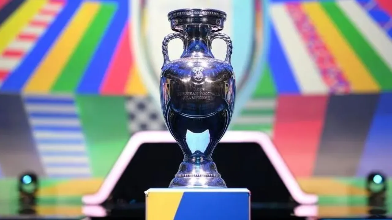 A imagem mostra o troféu da Eurocopa (UEFA European Championship Trophy), feito em prata, exposto sobre um pedestal colorido com fundo vibrante e moderno