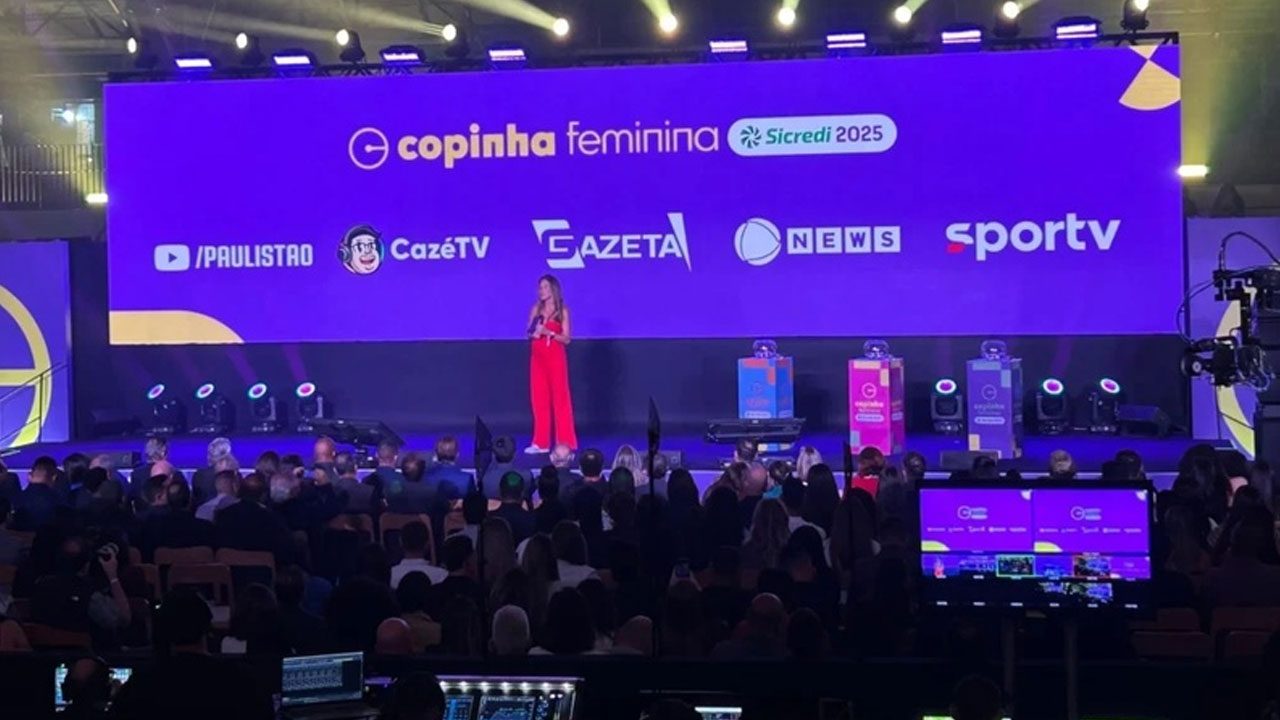 Mulher de macacão vermelho no palco de evento, com tela de fundo exibindo a marca Copinha Feminina Sicredi 2025 e logos de mídia