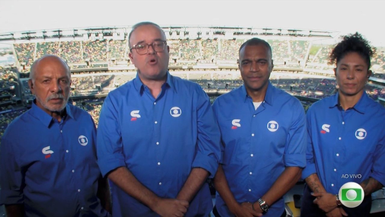 Everaldo Marques e equipe da Globo, de camisas azuis, na cabine de transmissão de Brasil x Haiti com estádio lotado ao fundo
