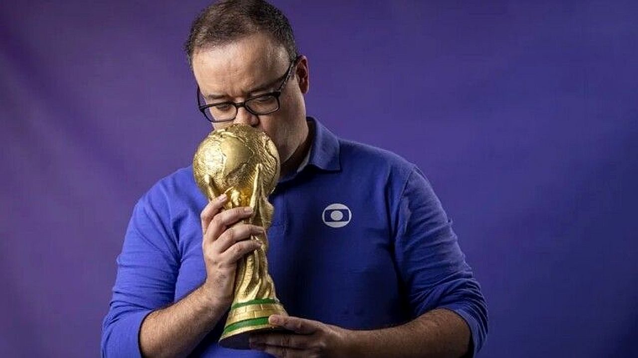 Homem de camisa da TV Globo beija réplica da taça da Copa do Mundo em fundo azul