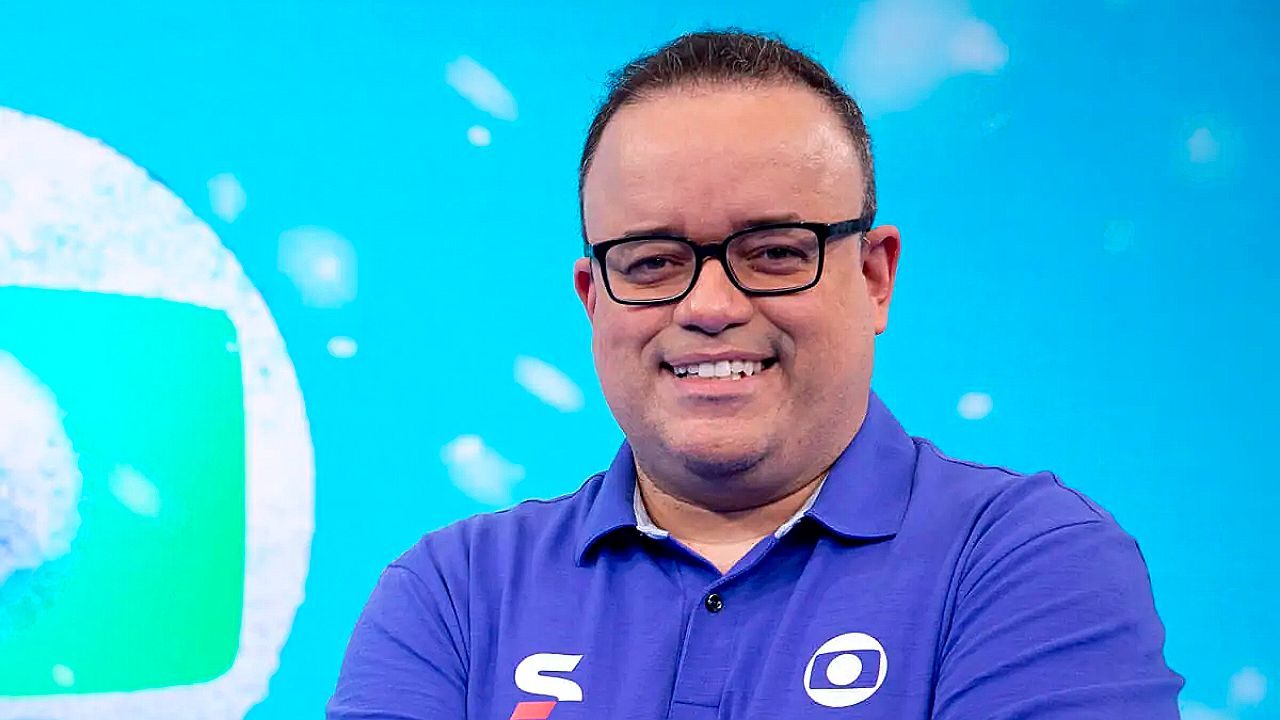 Homem de óculos sorri vestindo camisa polo da Globo em estúdio com fundo azul da emissora