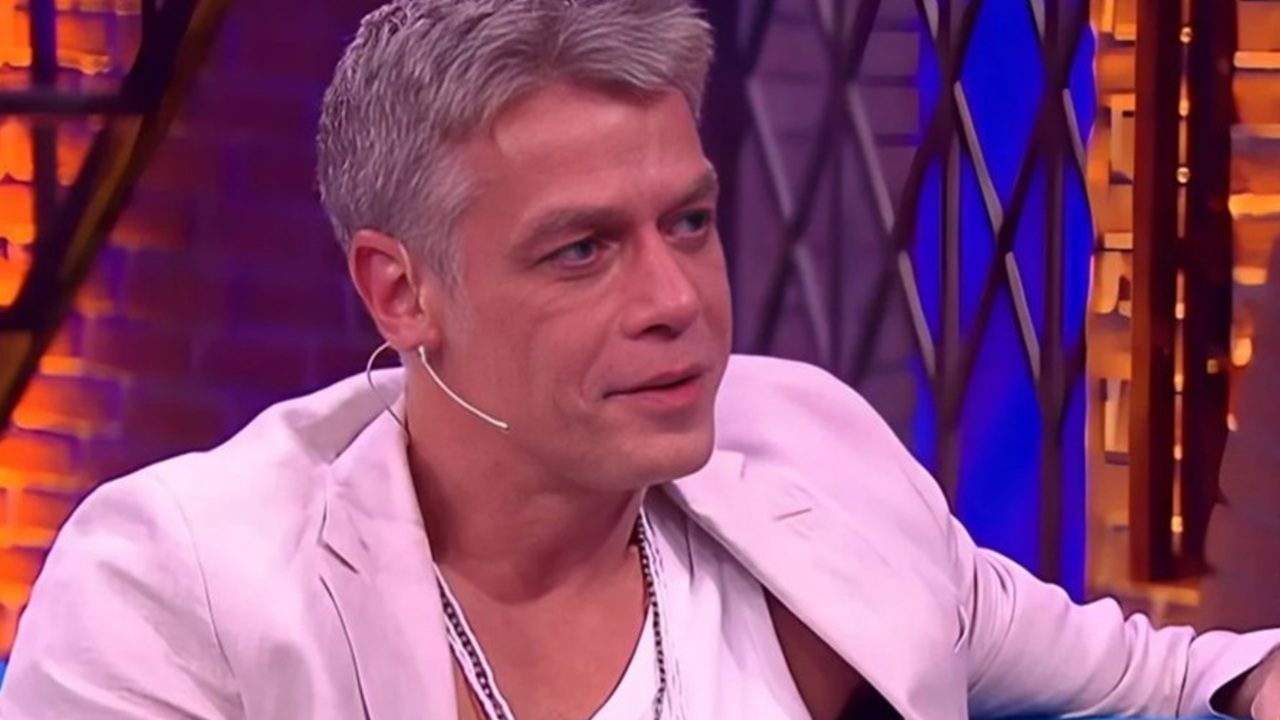 Fabio Assunção fala usando microfone de lapela em estúdio colorido com luzes roxas e alaranjadas ao fundo