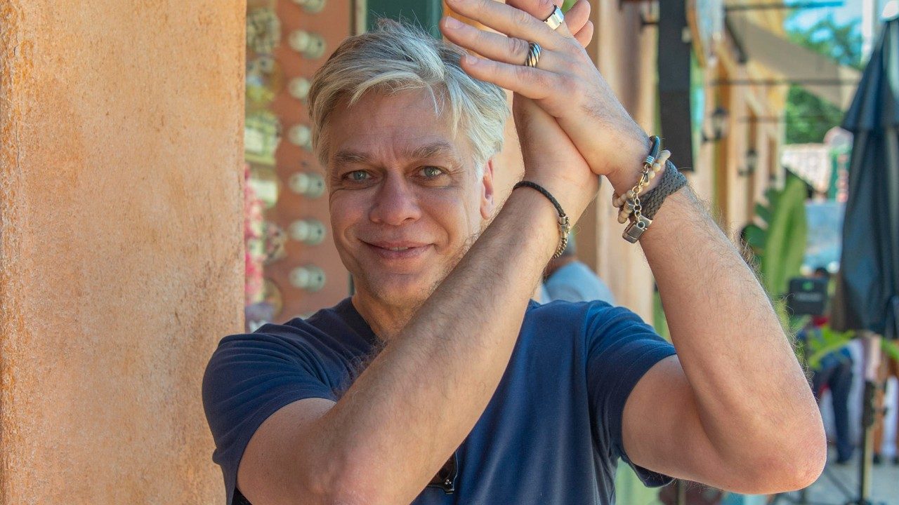 Fabio Assunção sorrindo com as mãos erguidas diante de prédio em área externa iluminada pelo sol