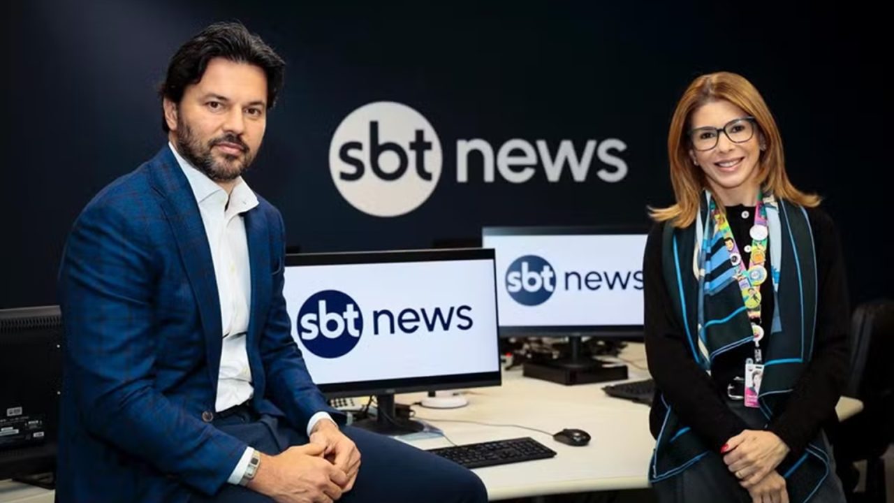 A imagem mostra Fábio Faria e Daniela Beyruti em um estúdio de redação do SBT News. Ao fundo, monitores e o logotipo do canal aparecem nas telas e na parede. Faria, de terno azul e camisa branca, está sentado sobre uma mesa, enquanto Daniela, de óculos e com um lenço colorido no pescoço, está em pé ao lado dele, ambos sorrindo levemente para a câmera