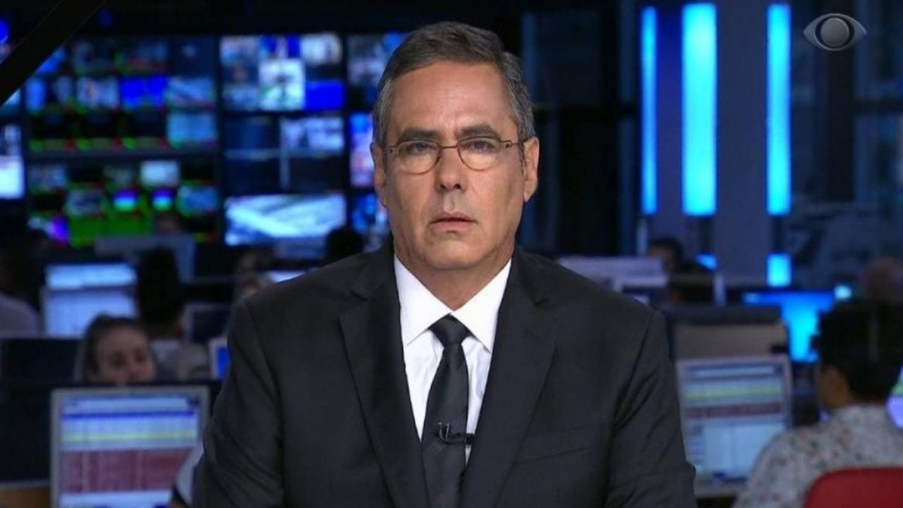 Homem de terno e gravata apresenta telejornal em estúdio de notícias, com monitores e equipe de produção ao fundo