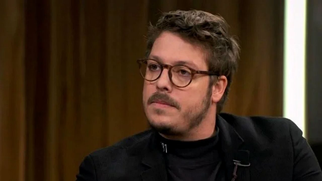 Homem de óculos e barba fala durante entrevista em programa de TV com fundo de madeira